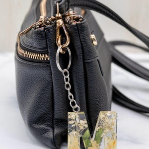 K&ouml;nnte beinhalten: Eine schwarze Ledertasche mit goldenen Rei&szlig;verschl&uuml;ssen und Beschl&auml;gen. Eine silberne Kette und ein Ring sind an einem Schl&uuml;sselanh&auml;nger mit dem Buchstaben "M" aus klarem Harz befestigt, der mit getrockneten Blumen gef&uuml;llt ist.