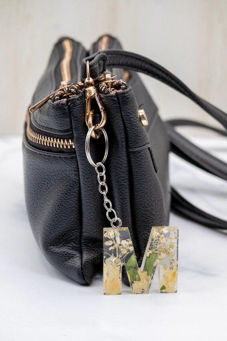 K&ouml;nnte beinhalten: Eine schwarze Ledertasche mit goldenen Rei&szlig;verschl&uuml;ssen und Beschl&auml;gen. Eine silberne Kette und ein Ring sind an einem Schl&uuml;sselanh&auml;nger mit dem Buchstaben "M" aus klarem Harz befestigt, der mit getrockneten Blumen gef&uuml;llt ist.