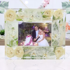 May include: A clear resin picture frame with white flowers and greenery embedded in the resin. A photo of a couple cutting a wedding cake is inside the frame.
