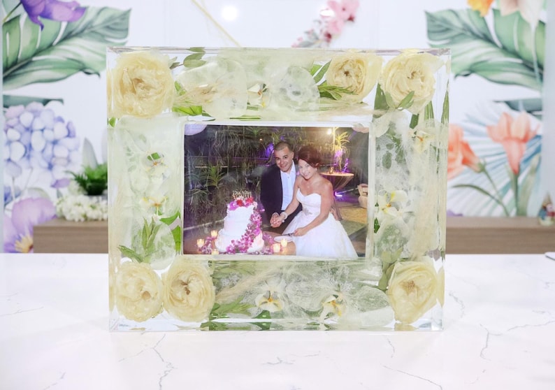 May include: A clear resin picture frame with white flowers and greenery embedded in the resin. A photo of a couple cutting a wedding cake is inside the frame.