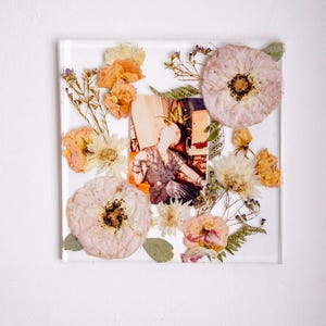 Pu&ograve; includere: Una cornice quadrata in resina trasparente con una fotografia vintage, circondata da fiori secchi nei toni del pesca, crema e giallo. La cornice &egrave; posta su uno sfondo bianco, creando un'esposizione delicata e artistica.