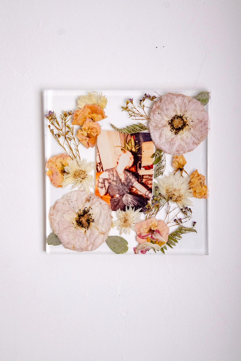 Pu&ograve; includere: Una cornice quadrata in resina trasparente con una fotografia vintage, circondata da fiori secchi nei toni del pesca, crema e giallo. La cornice &egrave; posta su uno sfondo bianco, creando un'esposizione delicata e artistica.