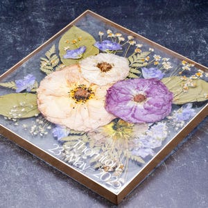 Pu&ograve; includere: Composizione floreale quadrata incorniciata con fiori secchi in resina trasparente. La composizione include fiori color crema e viola, foglie verdi e piccoli fiori blu. Il testo "Thornton 23 May 2025" &egrave; visibile.