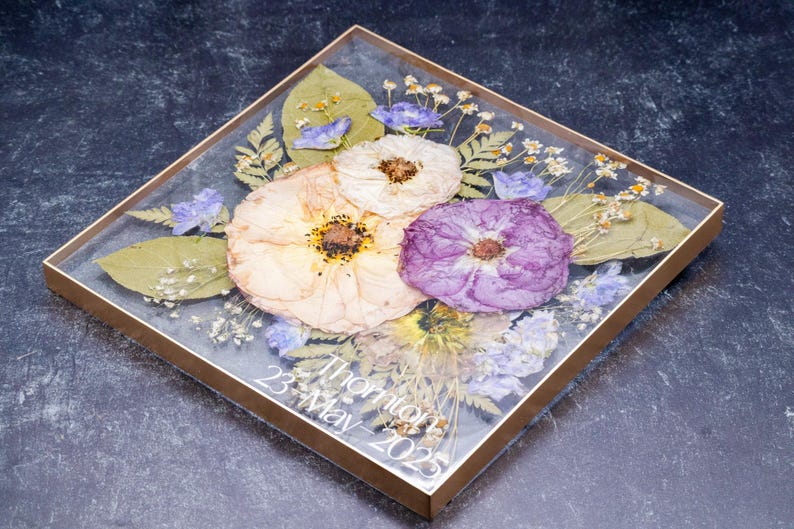Pu&ograve; includere: Composizione floreale quadrata incorniciata con fiori secchi in resina trasparente. La composizione include fiori color crema e viola, foglie verdi e piccoli fiori blu. Il testo "Thornton 23 May 2025" &egrave; visibile.