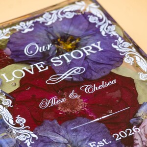 May include: A rectangular resin plaque featuring pressed purple and deep red flowers embedded within the clear material. The surface displays white printed text reading Our LOVE STORY, Alex & Chelsea, and Est. 2026, framed by a white decorative border.