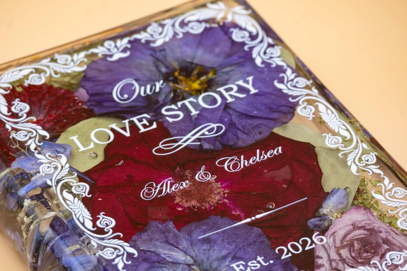 May include: A rectangular resin plaque featuring pressed purple and deep red flowers embedded within the clear material. The surface displays white printed text reading Our LOVE STORY, Alex & Chelsea, and Est. 2026, framed by a white decorative border.