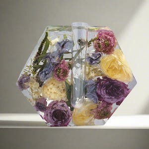 K&ouml;nnte beinhalten: Eine sechseckige, transparente Harzvase mit einem Glasrohr in der Mitte, gef&uuml;llt mit getrockneten Blumen in Violett-, Gelb- und Wei&szlig;t&ouml;nen.