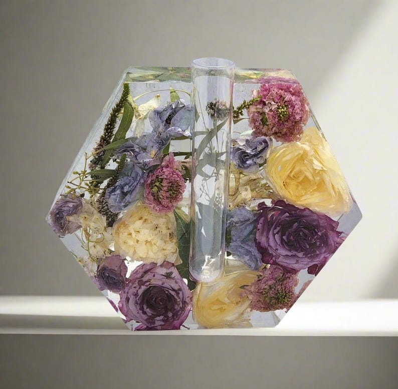 K&ouml;nnte beinhalten: Eine sechseckige, transparente Harzvase mit einem Glasrohr in der Mitte, gef&uuml;llt mit getrockneten Blumen in Violett-, Gelb- und Wei&szlig;t&ouml;nen.