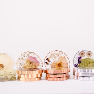 K&ouml;nnte beinhalten: Vier dekorative Untersetzer aus Kunstharz mit getrockneten Blumen. Jeder Untersetzer steht auf einem Stapel Metallringe in Gold-, Kupfer- und Silbert&ouml;nen. Die Untersetzer zeigen verschiedene Blumenarrangements.
