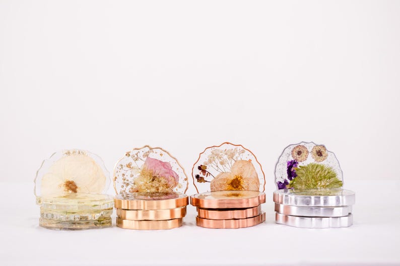 K&ouml;nnte beinhalten: Vier dekorative Untersetzer aus Kunstharz mit getrockneten Blumen. Jeder Untersetzer steht auf einem Stapel Metallringe in Gold-, Kupfer- und Silbert&ouml;nen. Die Untersetzer zeigen verschiedene Blumenarrangements.