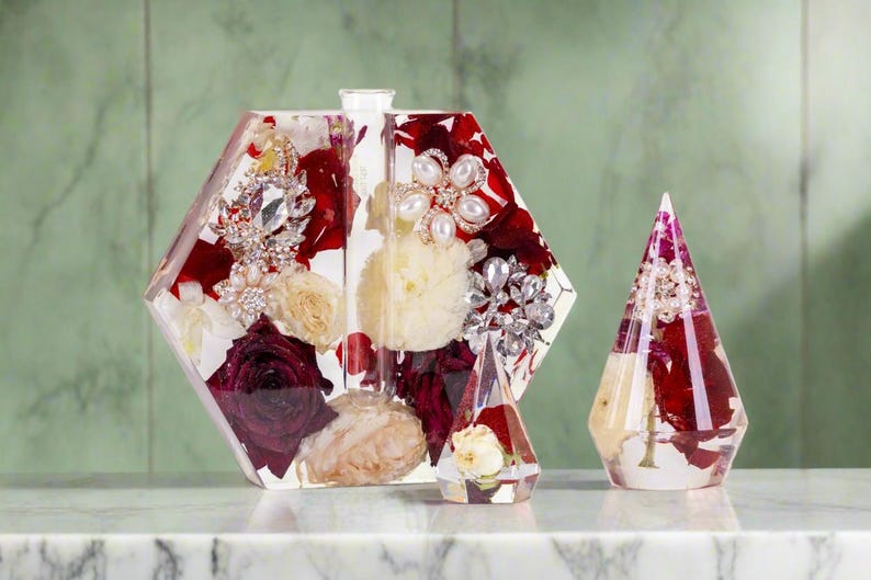 K&ouml;nnte beinhalten: Drei transparente Acrylbeh&auml;lter mit roten und wei&szlig;en Blumen und Perlen- und Strasssteinen. Die Beh&auml;lter sind in Form eines Sechsecks, eines Kegels und einer Pyramide.