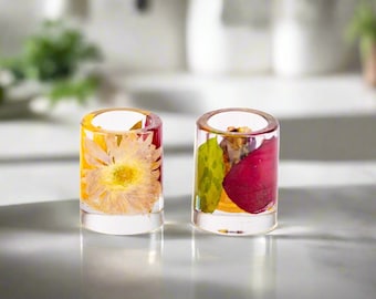 Könnte beinhalten: Zwei klare Kerzenhalter aus Kunstharz mit getrockneten Blumen und Blättern. Einer hat eine gelbe Blume, der andere ein rotes Blatt und andere Pflanzen. Die zylindrischen Halter stehen auf einer reflektierenden Oberfläche.