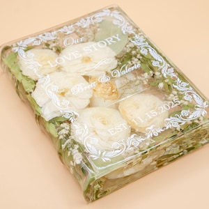 May include: A rectangular clear resin block containing preserved white flowers and green foliage. The surface features white printed text reading Our LOVE STORY, Brett & Valerie, and EST. 11.15.2025, framed by a decorative white filigree border.