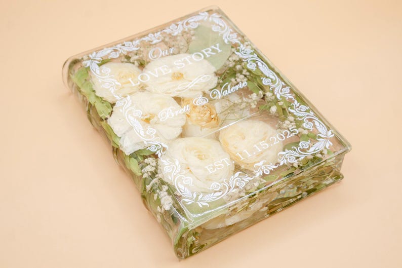 May include: A rectangular clear resin block containing preserved white flowers and green foliage. The surface features white printed text reading Our LOVE STORY, Brett & Valerie, and EST. 11.15.2025, framed by a decorative white filigree border.