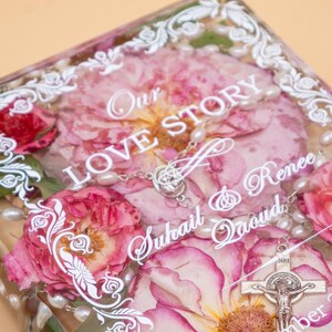 May include: A rectangular clear resin block containing pressed pink roses and a white pearl rosary. White text reads Our LOVE STORY, Suhail & Renee Qaoud, and a date. A silver crucifix is visible at the bottom right, framed by a white decorative border.