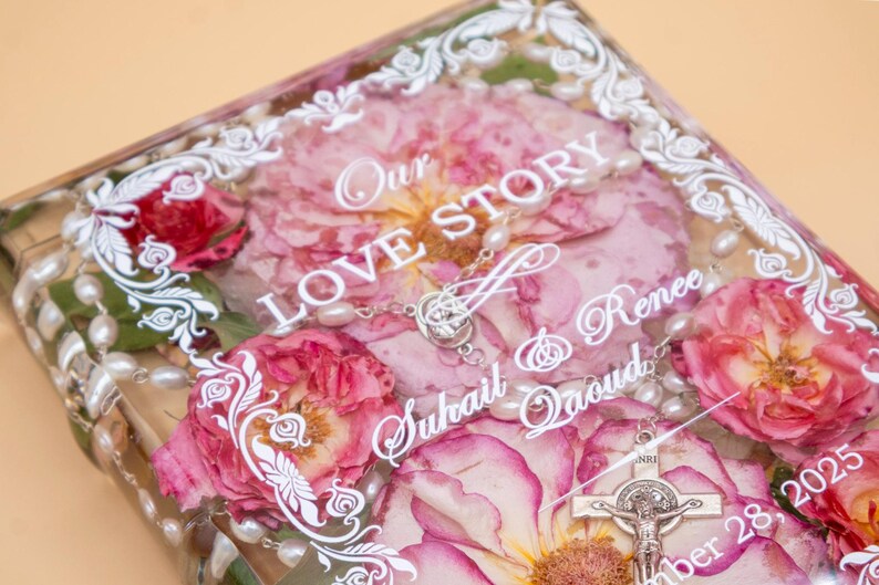 May include: A rectangular clear resin block containing pressed pink roses and a white pearl rosary. White text reads Our LOVE STORY, Suhail & Renee Qaoud, and a date. A silver crucifix is visible at the bottom right, framed by a white decorative border.