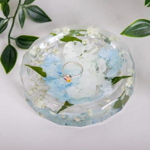 May include: A clear, faceted resin trinket dish containing embedded white and light blue pressed flowers and small white baby's breath blossoms. A thin, silver-toned metal ring with a small, multicolored iridescent stone sits in the center.