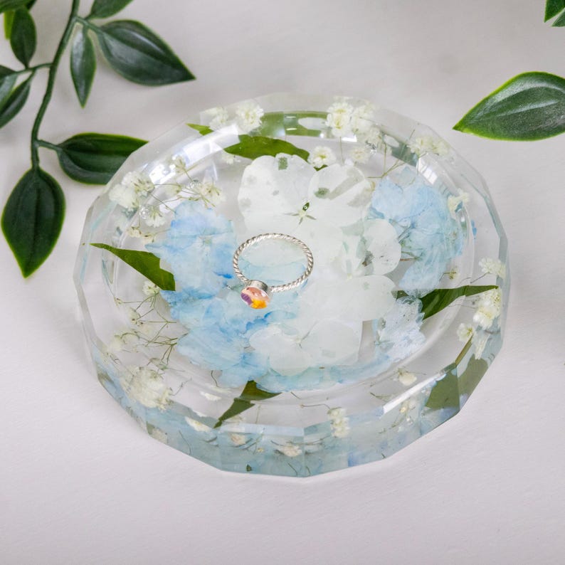 May include: A clear, faceted resin trinket dish containing embedded white and light blue pressed flowers and small white baby's breath blossoms. A thin, silver-toned metal ring with a small, multicolored iridescent stone sits in the center.