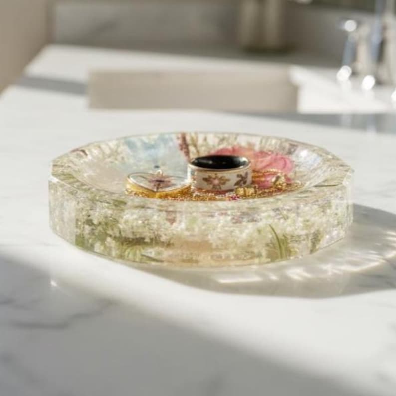 May include: A round, clear resin trinket dish embedded with small white flowers and pink petals. Inside the dish sits a gold-toned ring with a dark center and a gold-toned heart-shaped pendant with a delicate floral design on its surface.