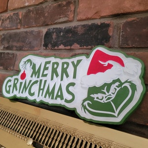 May include: A wooden sign with the words "MERRY GRINCHMAS" in white and green, featuring the Grinch wearing a Santa hat. A red ornament hangs from the letter "Y". The sign is placed in front of a brick wall.