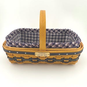 May include: Rectangular Longaberger Gathering Basket with a wooden handle. The basket is woven with a dark brown material and features a navy blue and white plaid fabric liner. A small tag on the side reads "Longaberger".