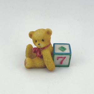 May include: A small, yellow teddy bear figurine with a red bow and a polka dot pattern. The bear sits next to a blue, green, and white block with the number 7 and a green diamond on it. The figurine is on a white background.