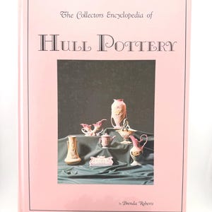 May include: A light pink hardcover book titled The Collectors Encyclopedia of Hull Pottery by Brenda Roberts. The cover features a central image of various pink and cream-colored ceramic vases and decorative pottery pieces.
