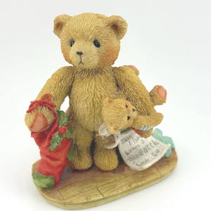 May include: A tan teddy bear figurine holding a red Christmas stocking with holly and berries. A smaller bear is held in its arms, and a note is attached. The figurine is on a wooden base.