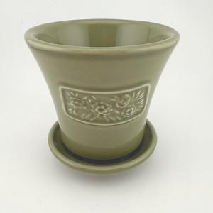May include: A sage green ceramic planter with a matching saucer. The planter has a flared shape and a rectangular embossed floral design. The planter is designed for indoor or outdoor use.