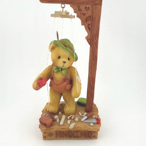 May include: A figurine of a teddy bear puppet wearing a green hat, brown overalls, and a green bow tie. The bear holds a red apple and stands on a wooden base with tools. A wooden frame with carved details supports the puppet strings.