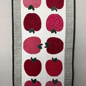 May include: A rectangular quilted wall hanging featuring eight red apple appliqués with green leaves on a white background. The apples have varying patterns. The border is a gray and white patterned fabric with a dark blue edge.