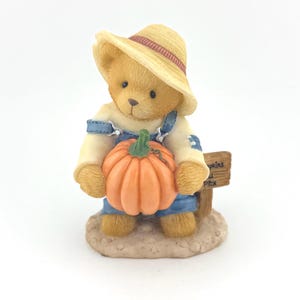 May include: A figurine of a teddy bear wearing a straw hat and denim overalls, holding an orange pumpkin. The bear stands in front of a small sign that reads "Pumpkins Pick". The figurine has a beige base.