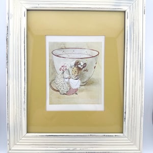May include: A framed print featuring a whimsical illustration of a mouse dressed in a bonnet and apron, standing near a teacup. The print is set against a yellow background and framed in a distressed white wooden frame, creating a vintage aesthetic.