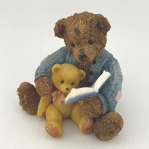 May include: A brown teddy bear figurine wearing a blue sweater, holding a book and embracing a smaller yellow teddy bear with a pink bow. The bears are sitting, and the book is open, with white pages.