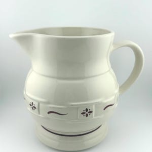 May include: Cream-colored ceramic pitcher with a curved handle and a spout. The pitcher features a decorative band with dark red floral and curved line accents. A classic piece for serving beverages.
