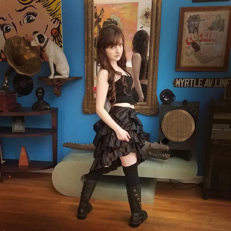 May include: A woman wearing a black corset top and a black and brown layered skirt. She is wearing black knee-high boots and has long dark hair. She is standing in a room with a blue wall and a mirror behind her. The text "MYRTLE AV. LINE" is visible on the wall.