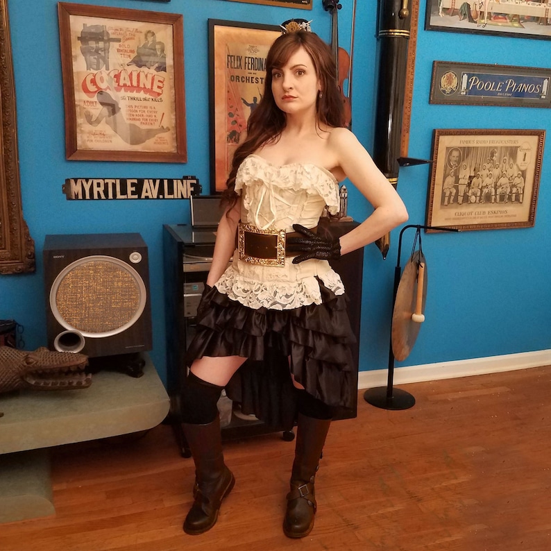 May include: A woman wearing a white lace corset with a gold belt, a black skirt with ruffles, and brown boots. She is standing in front of a blue wall with various framed posters and a large speaker.