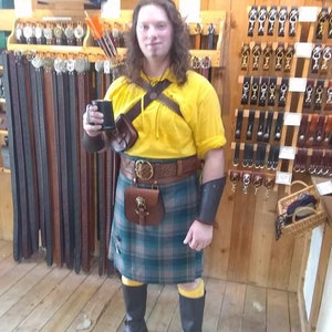 May include: A person wearing a yellow shirt, a green and black kilt, and black boots. They are holding a black mug in their right hand. The person is standing in front of a wall with shelves of leather goods.