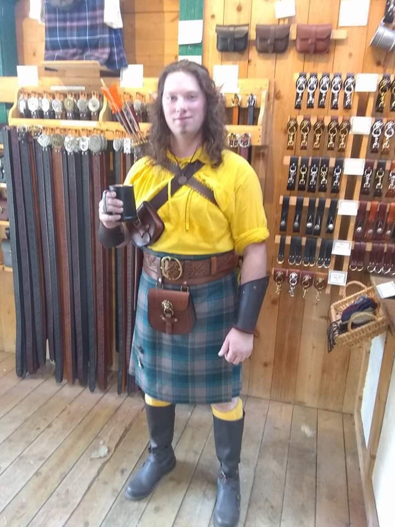 May include: A person wearing a yellow shirt, a green and black kilt, and black boots. They are holding a black mug in their right hand. The person is standing in front of a wall with shelves of leather goods.