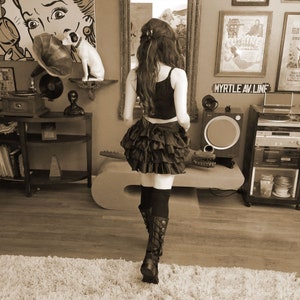 May include: A woman wearing a black and white ruffled skirt and black boots stands in a room with a vintage record player, a framed picture of a woman, and a framed movie poster. The poster reads "Myrtle Av. Line".