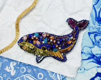 Op de afbeelding: Een broche in de vorm van een walvis, versierd met kralen in blauw, paars en goud. De walvis is gemaakt van pailletten, kralen en draad. Het is een handgemaakt sieraad.