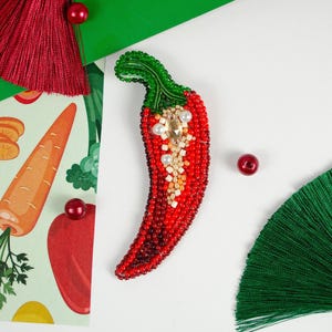 May include: A red beaded chili pepper brooch with a green stem and white pearl accents. The brooch is on a white background.