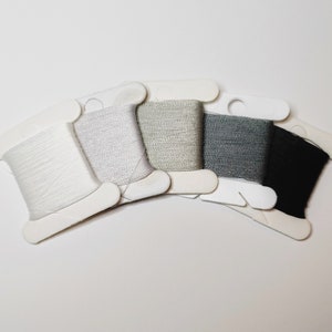 Puede incluir: Cinco carretes de hilo en tonos de blanco, gris y negro están dispuestos en forma de abanico. Cada carrete está montado en una tarjeta blanca. Los colores del hilo van del blanco brillante al negro carbón.