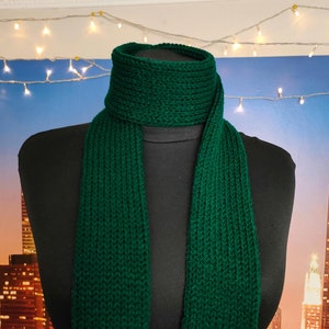 May include: A dark green knitted scarf with a ribbed texture. The scarf is displayed on a black mannequin against a backdrop of a city skyline.