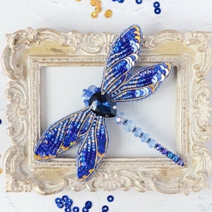 Può includere: Un broche a forma di libellula blu e dorata fatto di paillettes e perline. Il broche è esposto in una cornice bianca decorata.