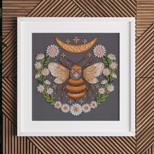 May include: A framed beaded artwork featuring a bee with a crescent moon and stars above it, surrounded by a wreath of white flowers on a gray background.
