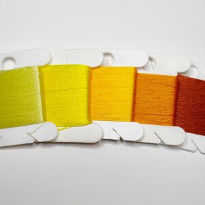 Puede incluir: Cinco carretes de hilo de bordar en un degradado de amarillo, naranja y rojo anaranjado. El hilo está cuidadosamente enrollado en bobinas de cartón blanco. Los colores van del amarillo claro al naranja rojizo oscuro.