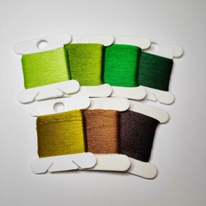 Puede incluir: Una colección de ocho carretes de hilo de bordar en varios tonos de verde y marrón. Los colores van desde el verde lima claro hasta el verde bosque oscuro, con tonos adicionales de oliva, beige y marrón oscuro. Cada carrete está montado en una tarjeta blanca.