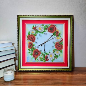 May include: A framed clock with a red background and a white clock face. The clock face is surrounded by a wreath of red poppies and white daisies. The clock hands are black and the numbers are Roman numerals.