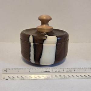 Może przedstawiać: Ręcznie wykonane drewniane pudełko z pokrywką. Pudełko jest ciemnobrązowe z pionowym białym paskiem. Pokrywka ma jasnobrązowy uchwyt. Pudełko ma średnicę około 10 cm.